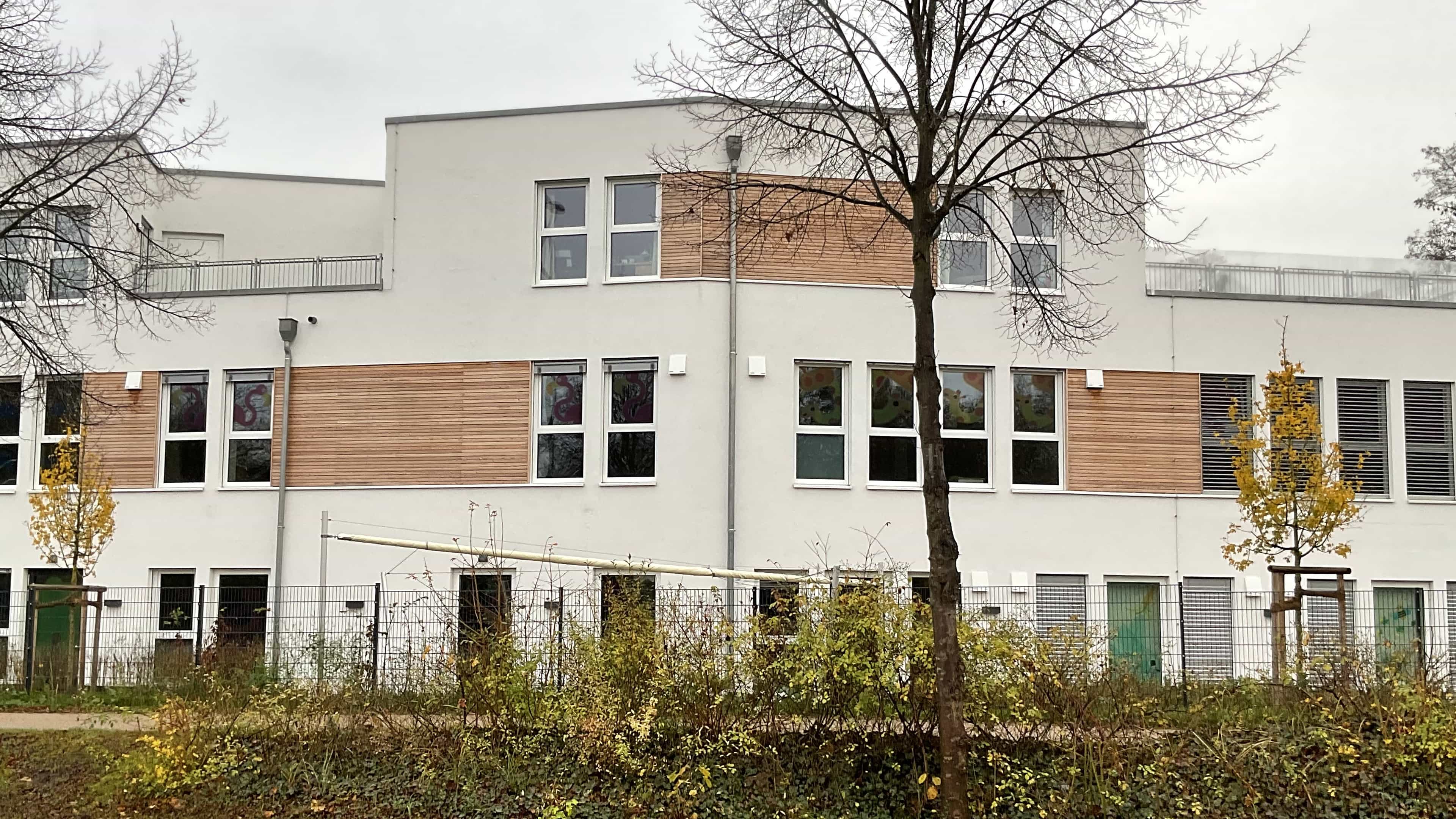 Weiße Fassade der Kita KD1 mit Holzverkleidung, Fensterreihen und Bäumen davor