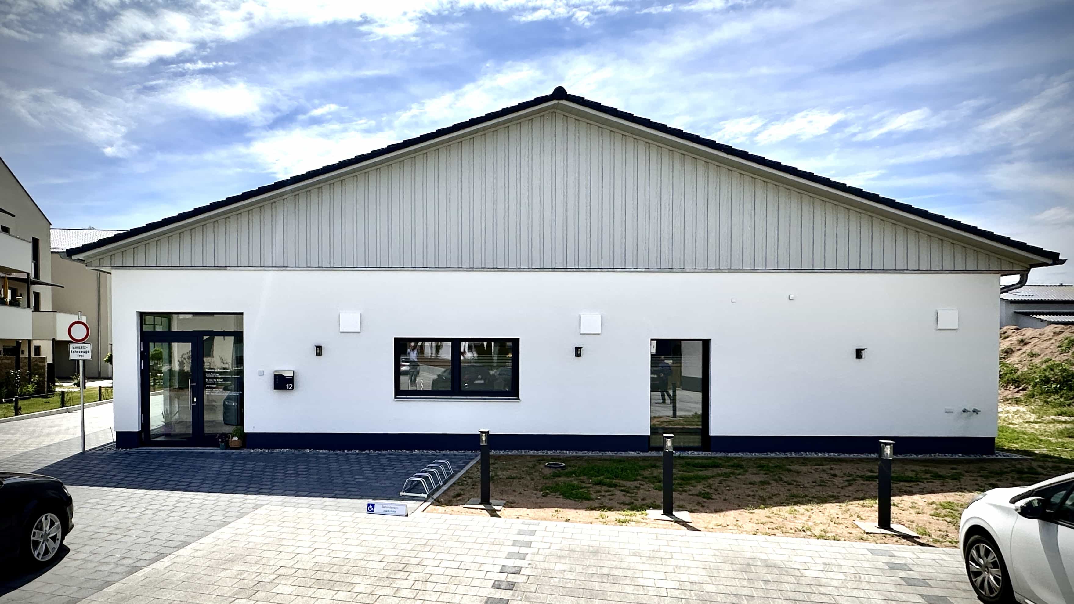 Eingangsseite des Praxisgebäudes Balzer mit Glastür, Satteldach und Parkplätzen