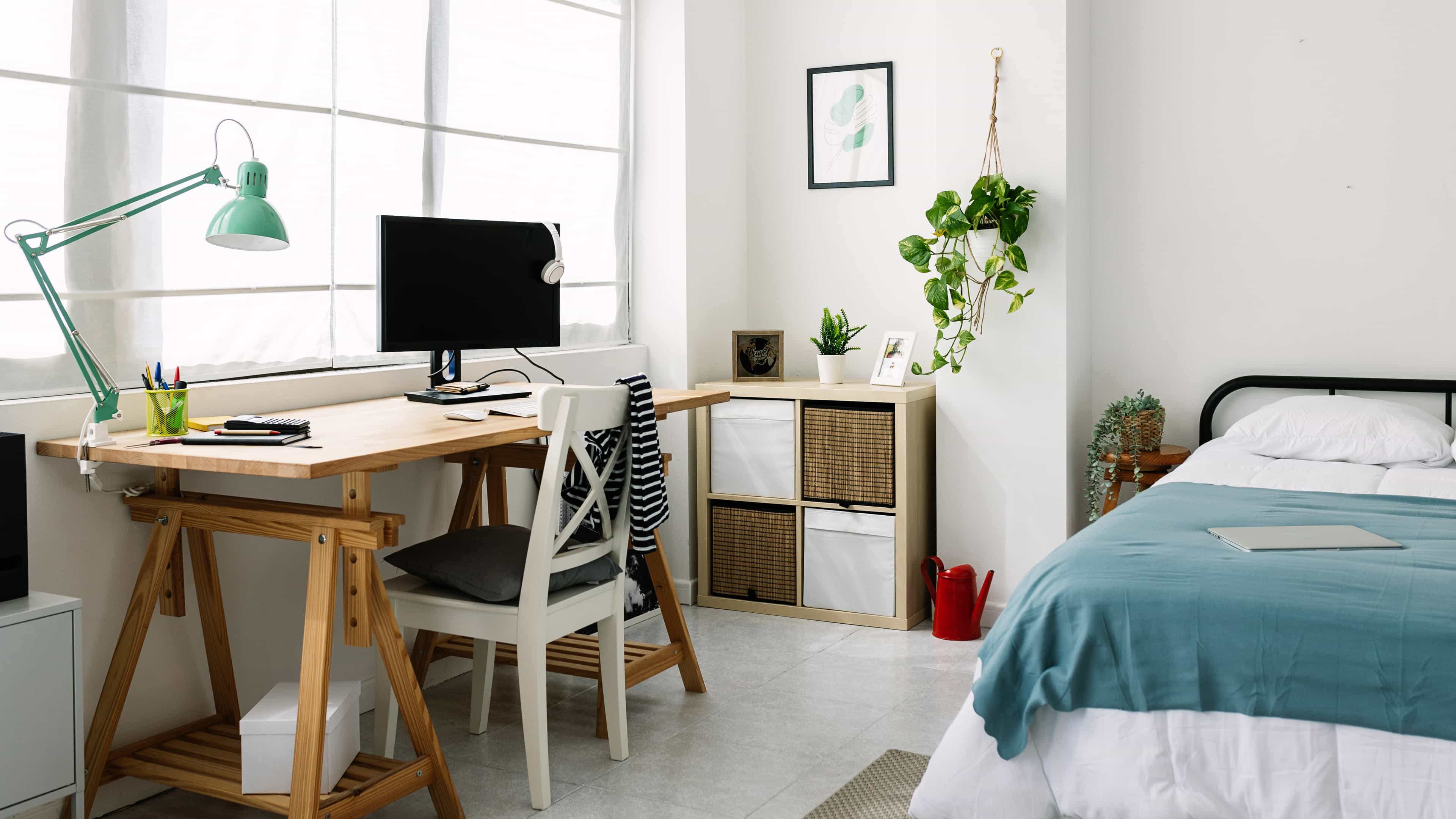 Helles Arbeitszimmer mit Schreibtisch, Stuhl, Computer und Lampe neben einem Bett mit blauer Decke.