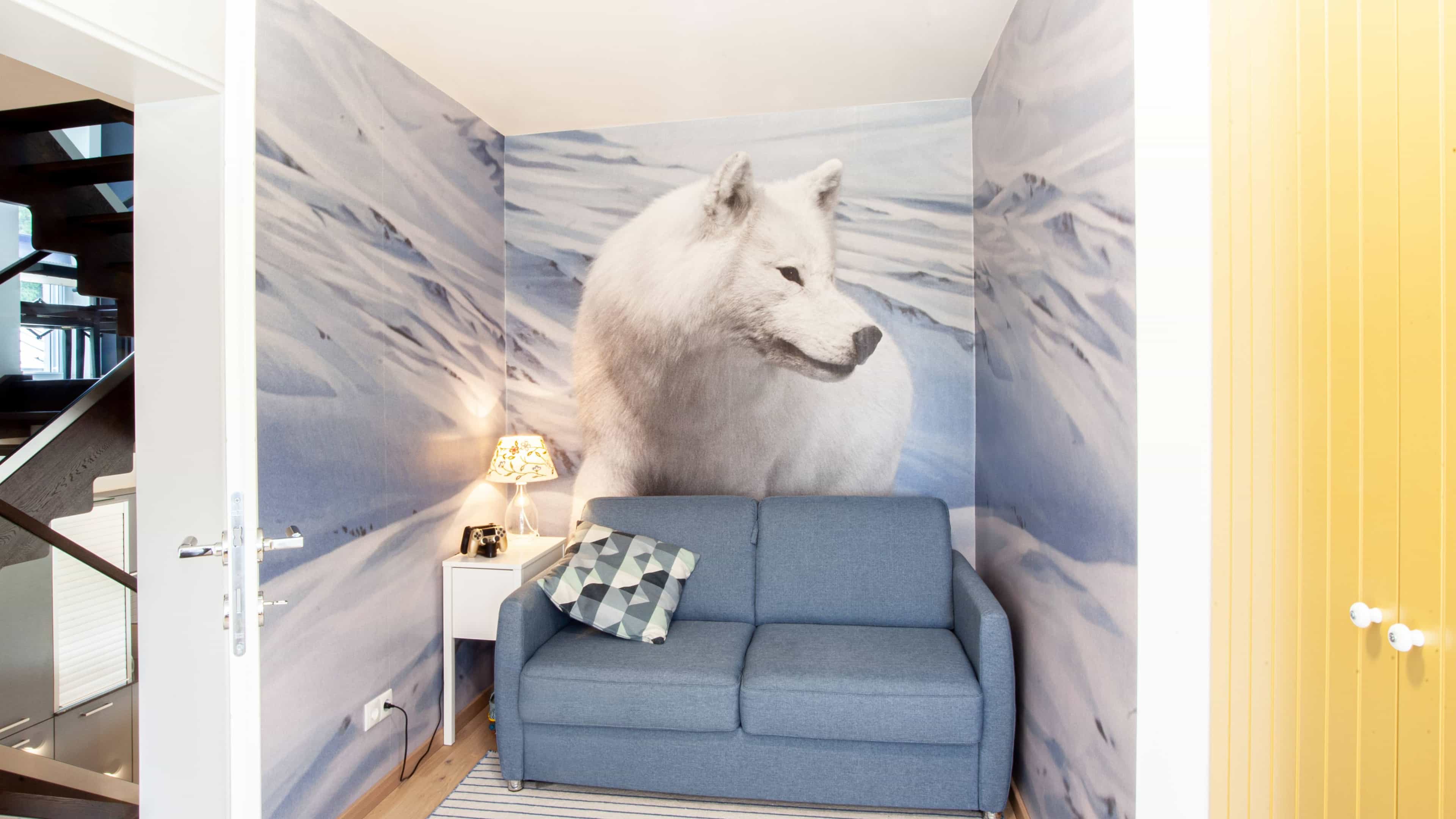 Kompaktes Wohnzimmer mit blauer Couch, einer Lampe und Wandbild eines weißen Wolfes auf schneebedecktem Hintergrund.