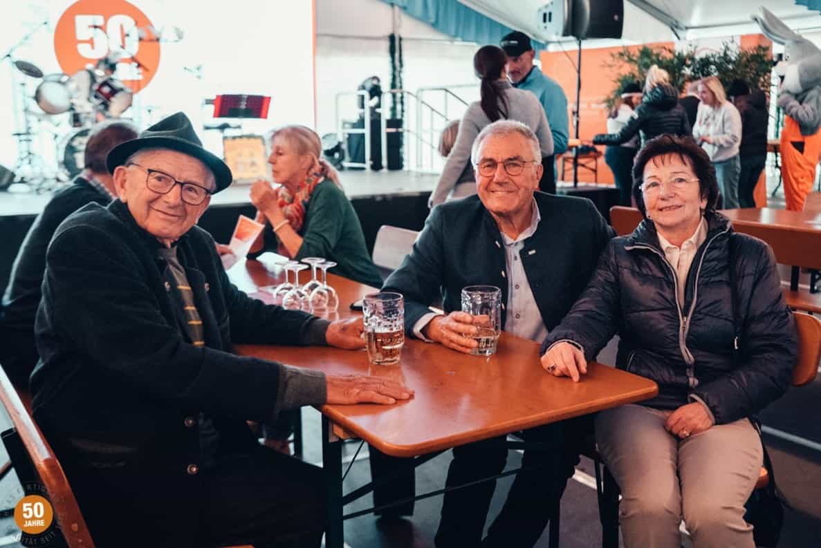 Ältere Menschen sitzen bei einem Fest an einem Tisch, halten Biergläser und lächeln; im Hintergrund Bühne mit Schlagzeug.