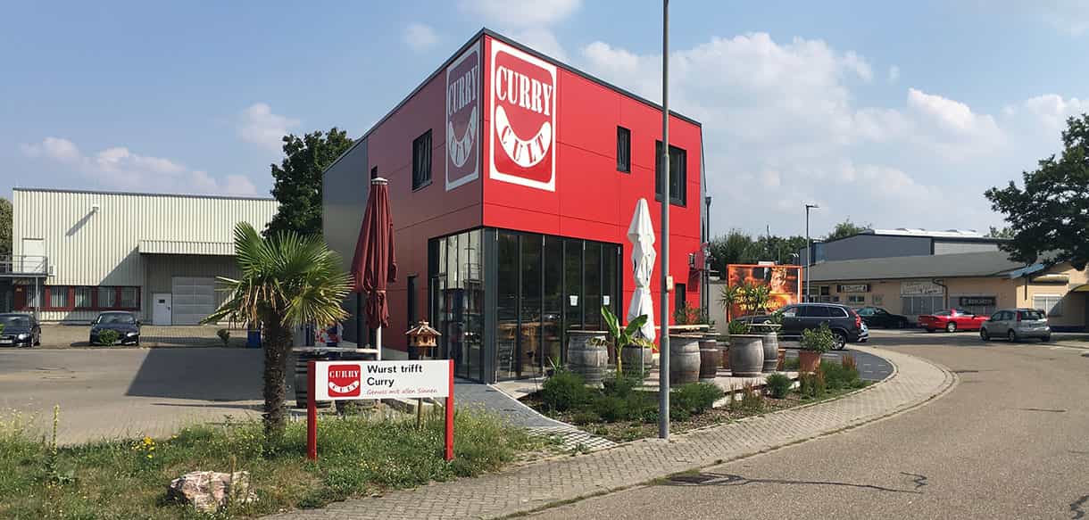 Dreieckiges Gebäude von Curry Cult mit roter Fassade und großflächiger Eingangsverglasung, Straßenansicht