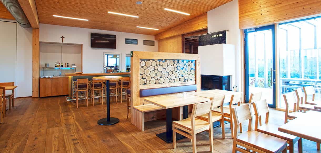 Gastraum des KWB-Ausstellungsgebäudes mit Holztischen, Theke und Holzscheitwand