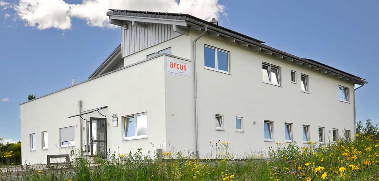Seitenansicht der Arcus Praxis für Physiotherapie mit Logo an der Fassade und Blumenwiese davor
