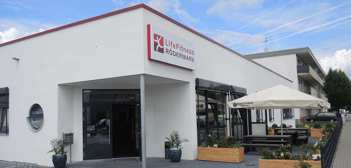 Eingang mit Vordach und Logo von Life Fitness Rödermark mit Terrasse und Sonnenschirm