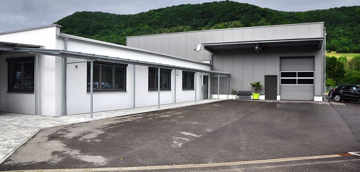 Betriebsgebäude von BC Sports in Deggingen mit Bürotrakt, Hallentor und Parkplatz vor bewaldeten Hügeln
