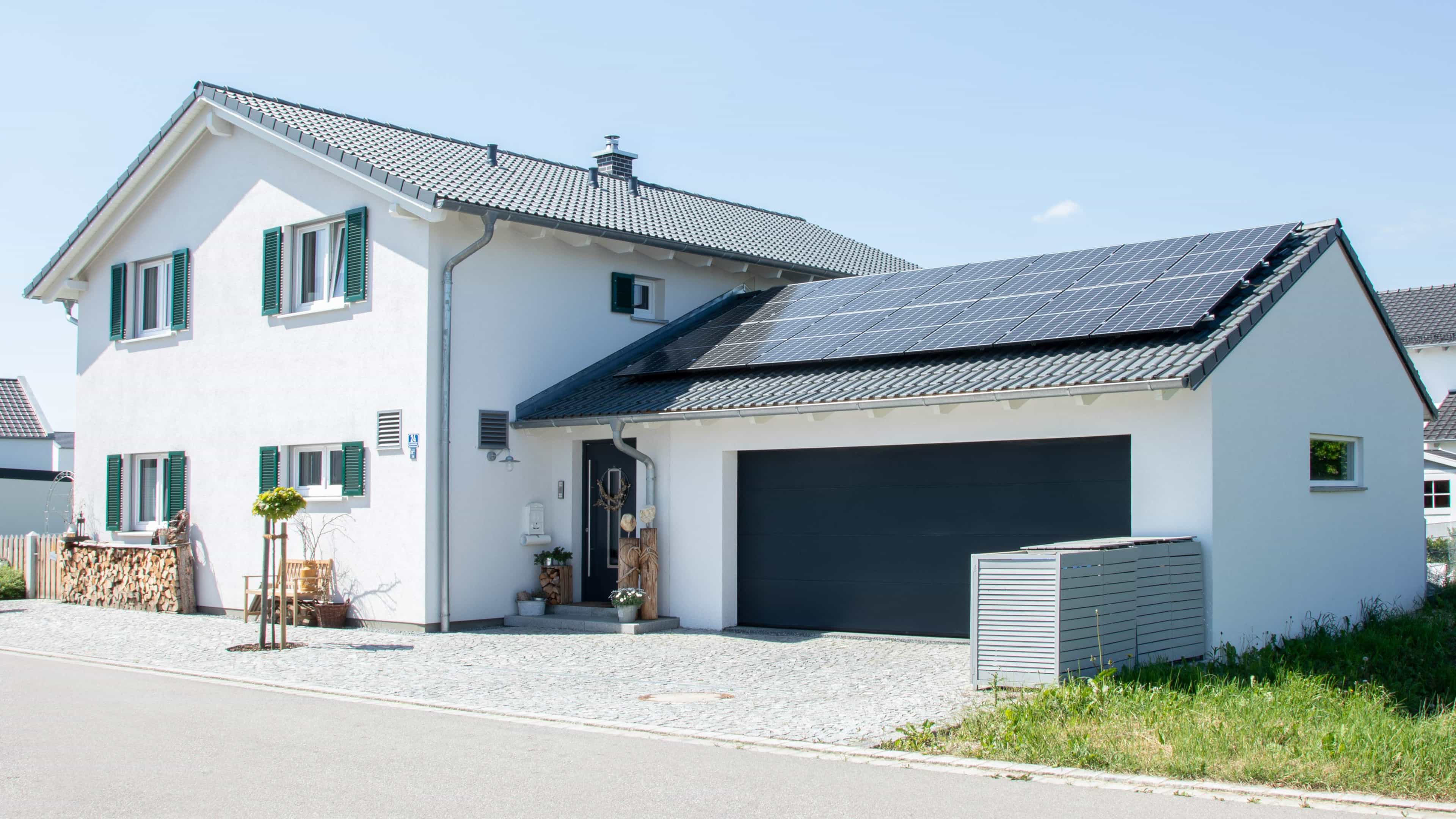 Modernes Einfamilienhaus mit weißer Fassade, Solarpanelen auf dem Dach, einer Garage und grünem Vorgarten an der Straße.