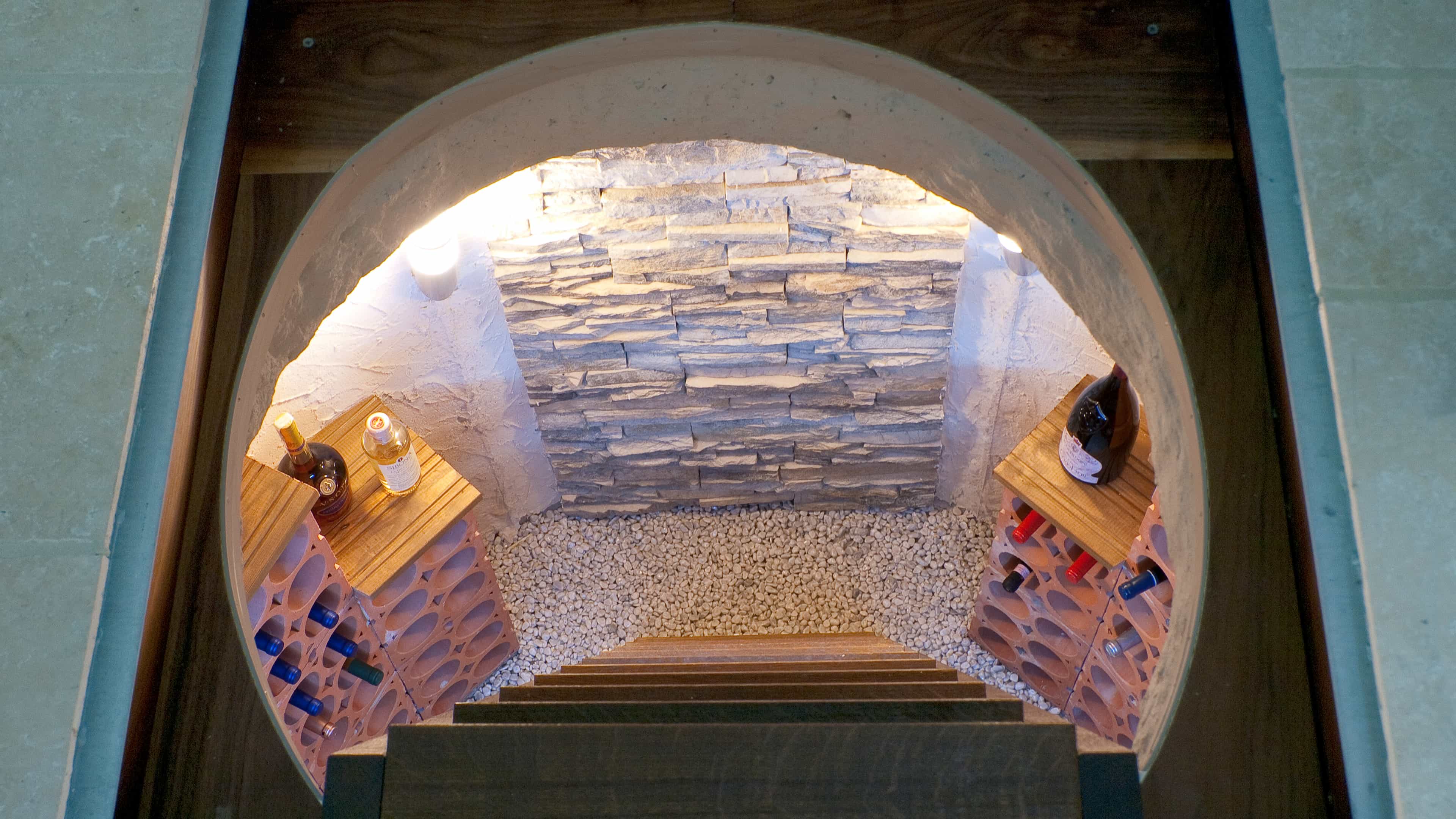 Blick in einen kleinen Weinkeller mit Holzböden und Steinwand, gefüllt mit Weinflaschen und einigen Flaschen auf Regalen.