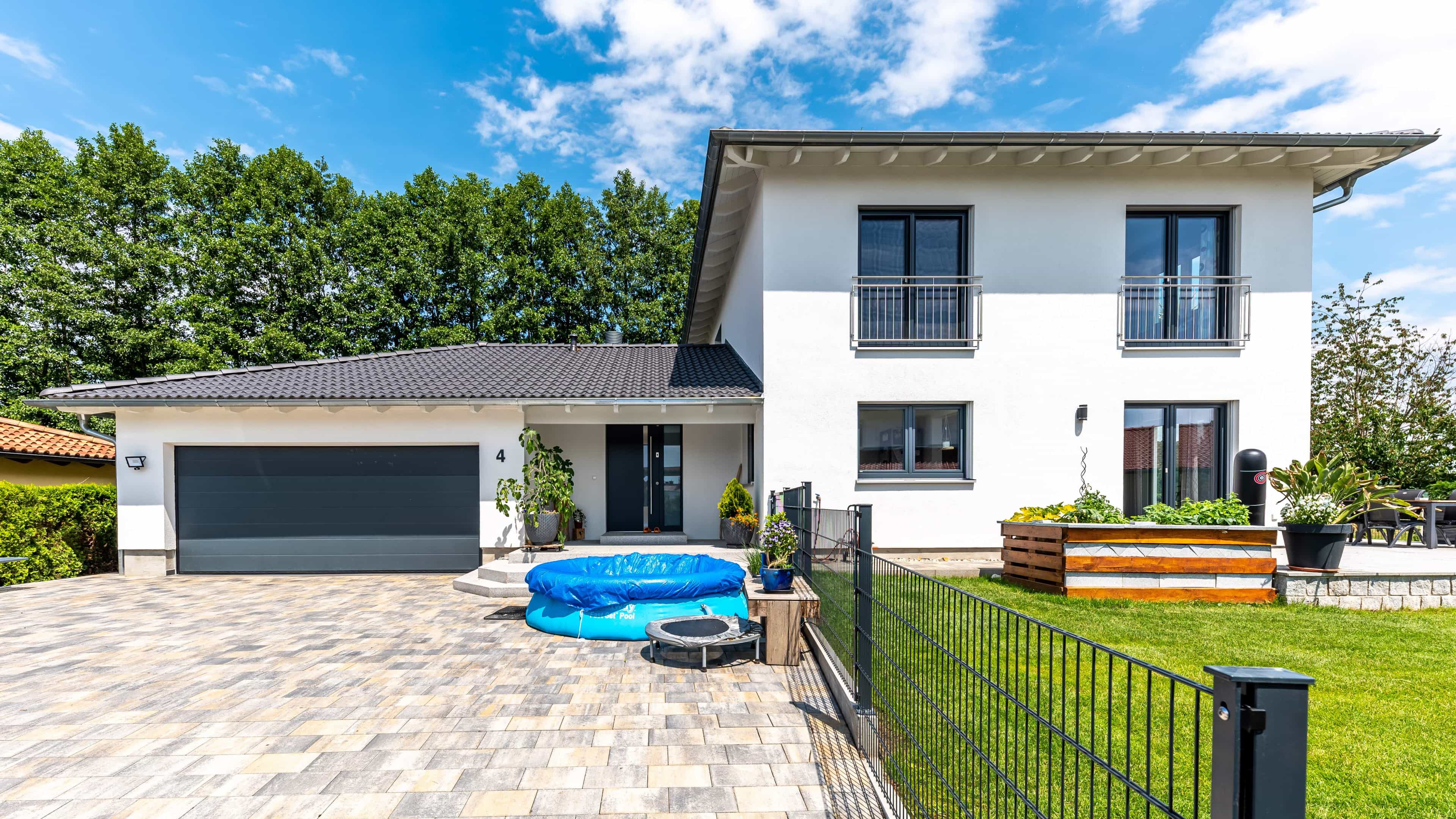 Modernes zweistöckiges Einfamilienhaus mit Garage, Pool im Garten und umgeben von Bäumen unter blauem Himmel.