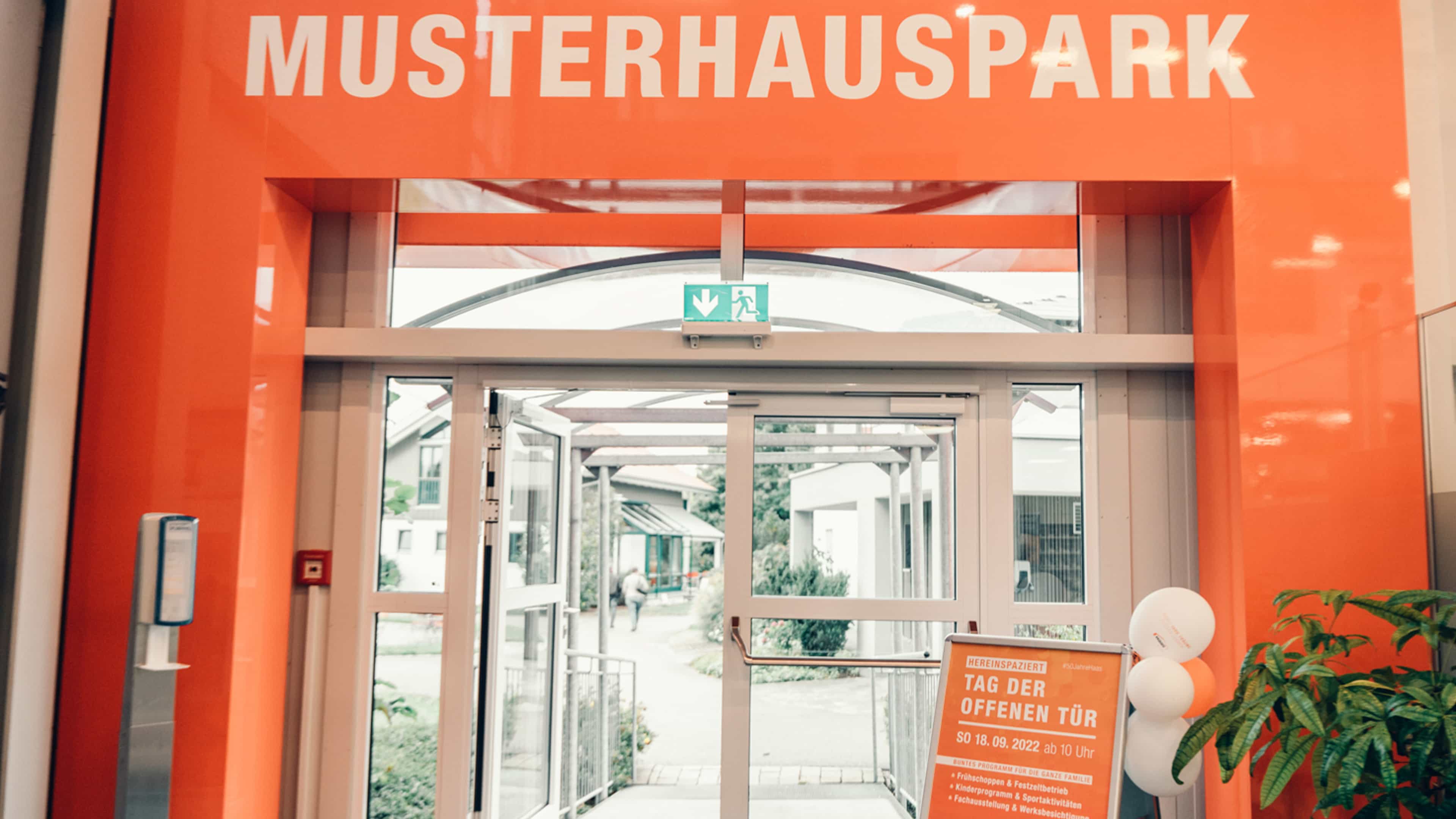 Eingang eines Musterhausparks mit Schild zum "Tag der offenen Tür" und Dekoration im Innenbereich.