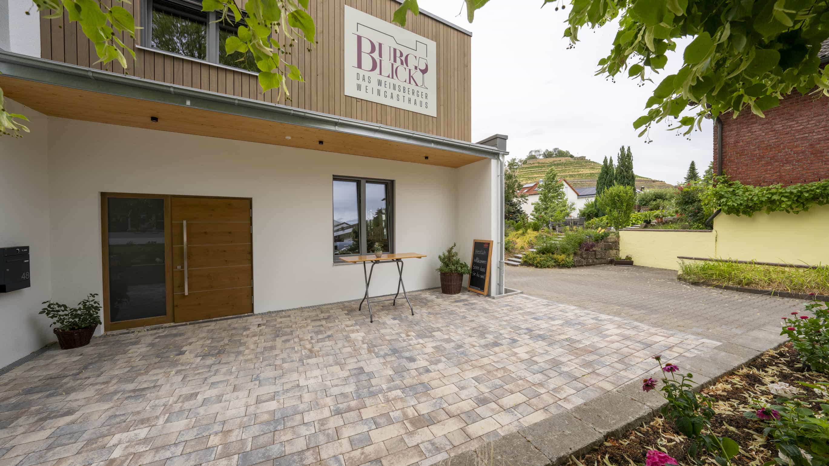 Eingang der Weinwirtschaft Burgblick mit Holztür, Schild und gepflastertem Vorplatz, Haas Gewerbebau