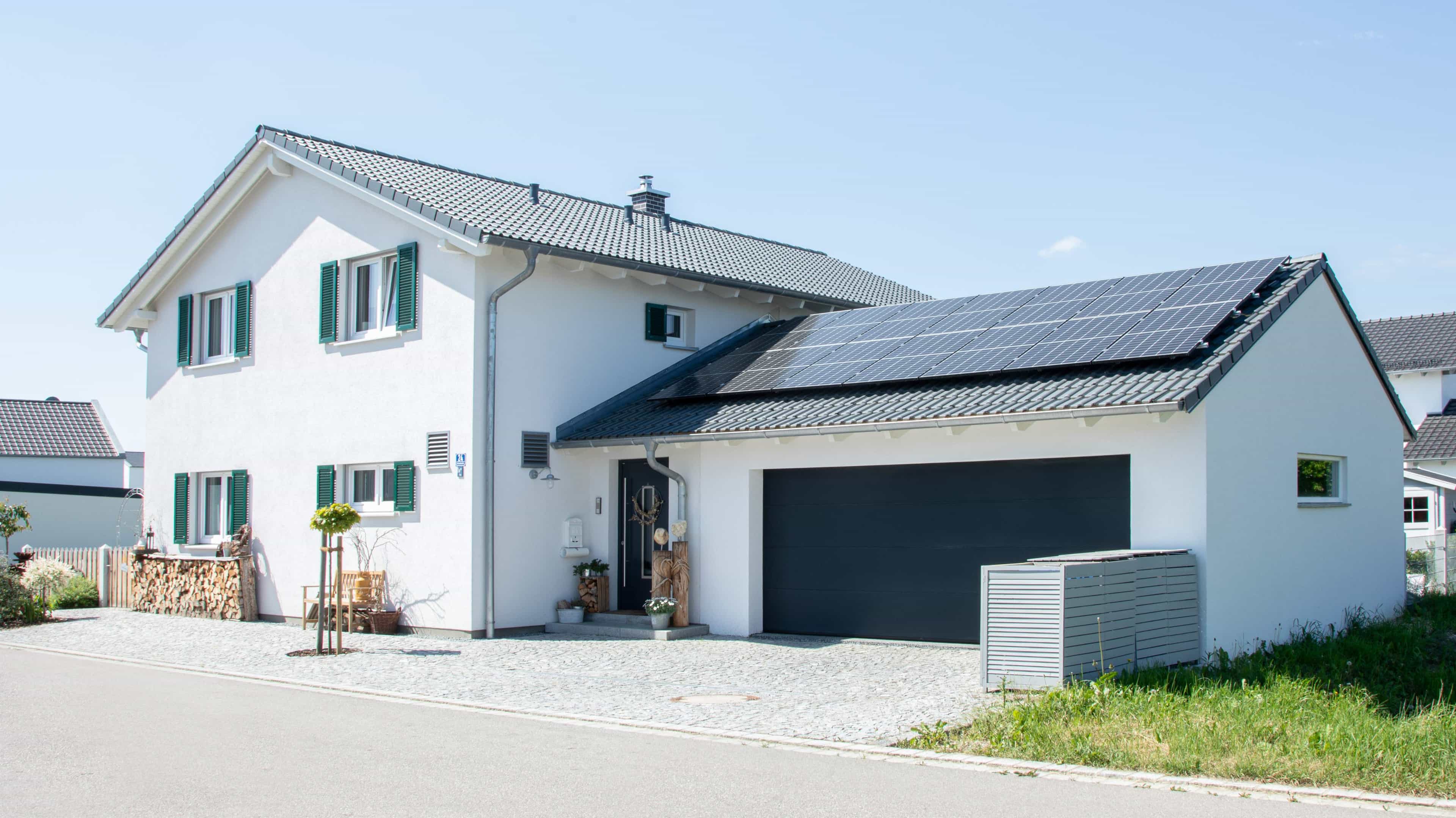 Modernes zweistöckiges Einfamilienhaus mit Solarmodulen auf dem Garagendach und grünen Fensterläden, sonniger Tag.