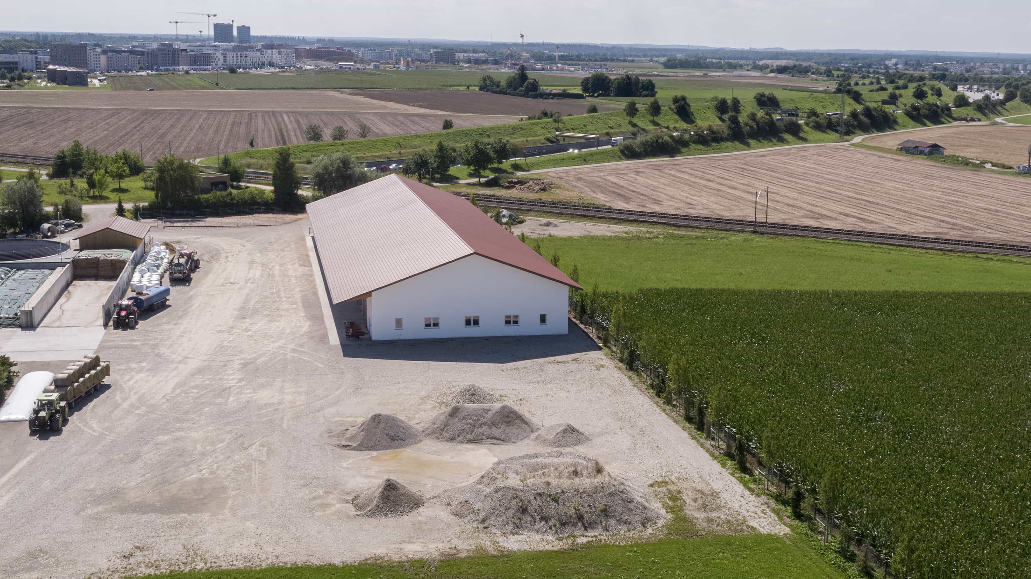Luftaufnahme der langgestreckten weißen Maschinenhalle mit rotem Dach zwischen Feldern