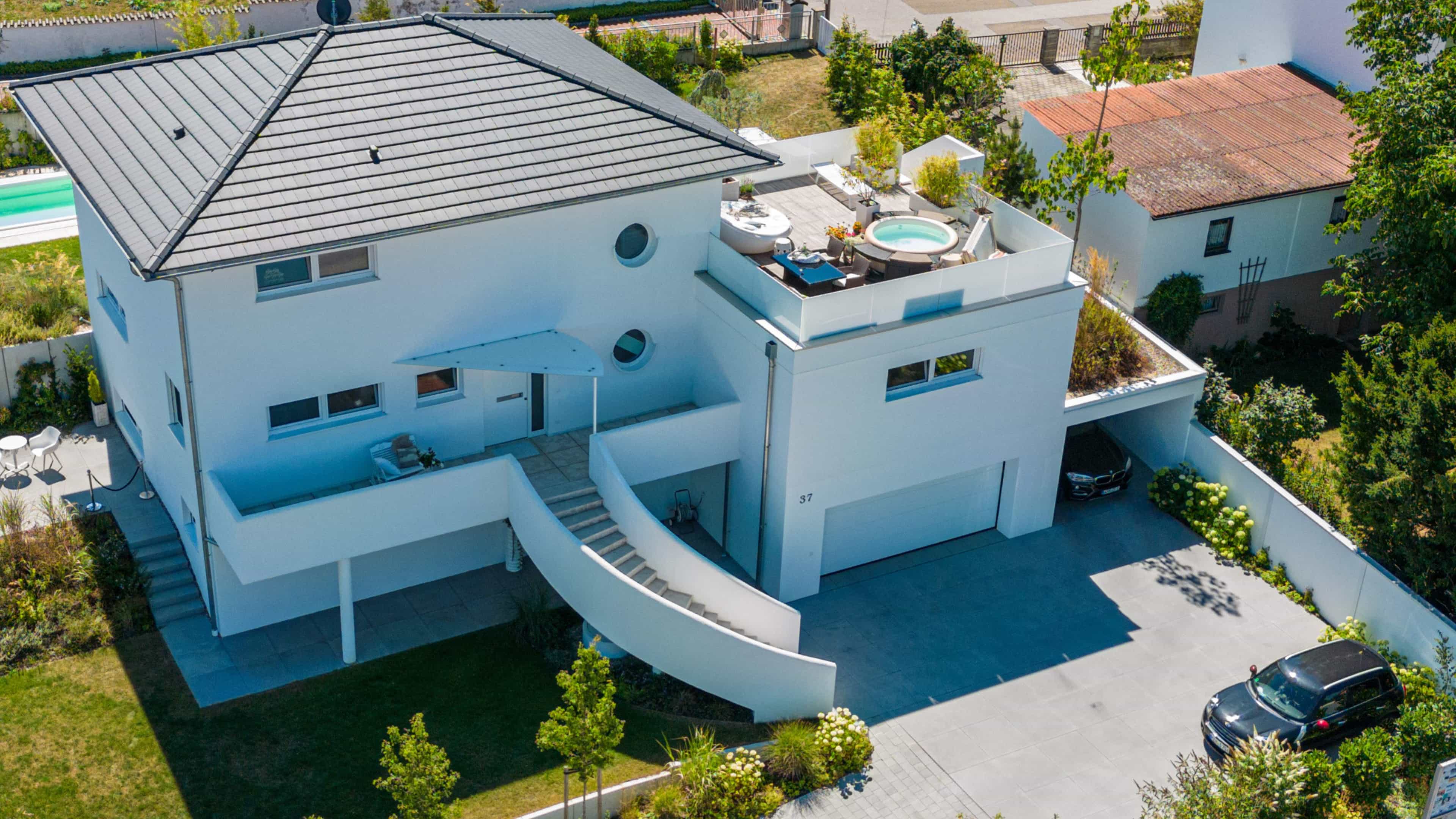 Modernes weißes Haus mit Dachterrasse, Whirlpool, geschwungener Außentreppe und geparkten Autos in einem gepflegten Garten.