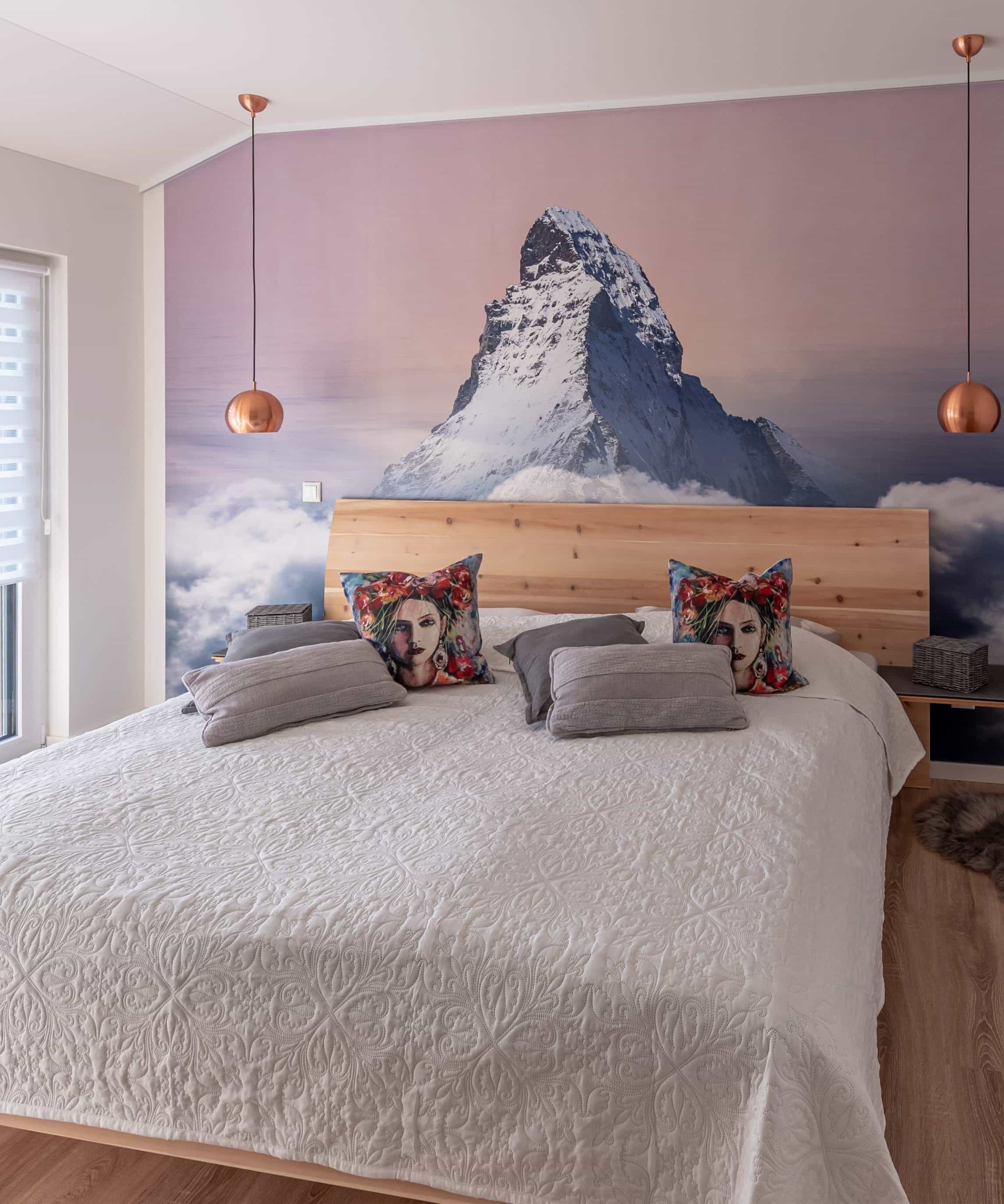 Modernes Schlafzimmer mit Bett, Wolldecken und Bergfototapete, durch große Fenster gut beleuchtet.