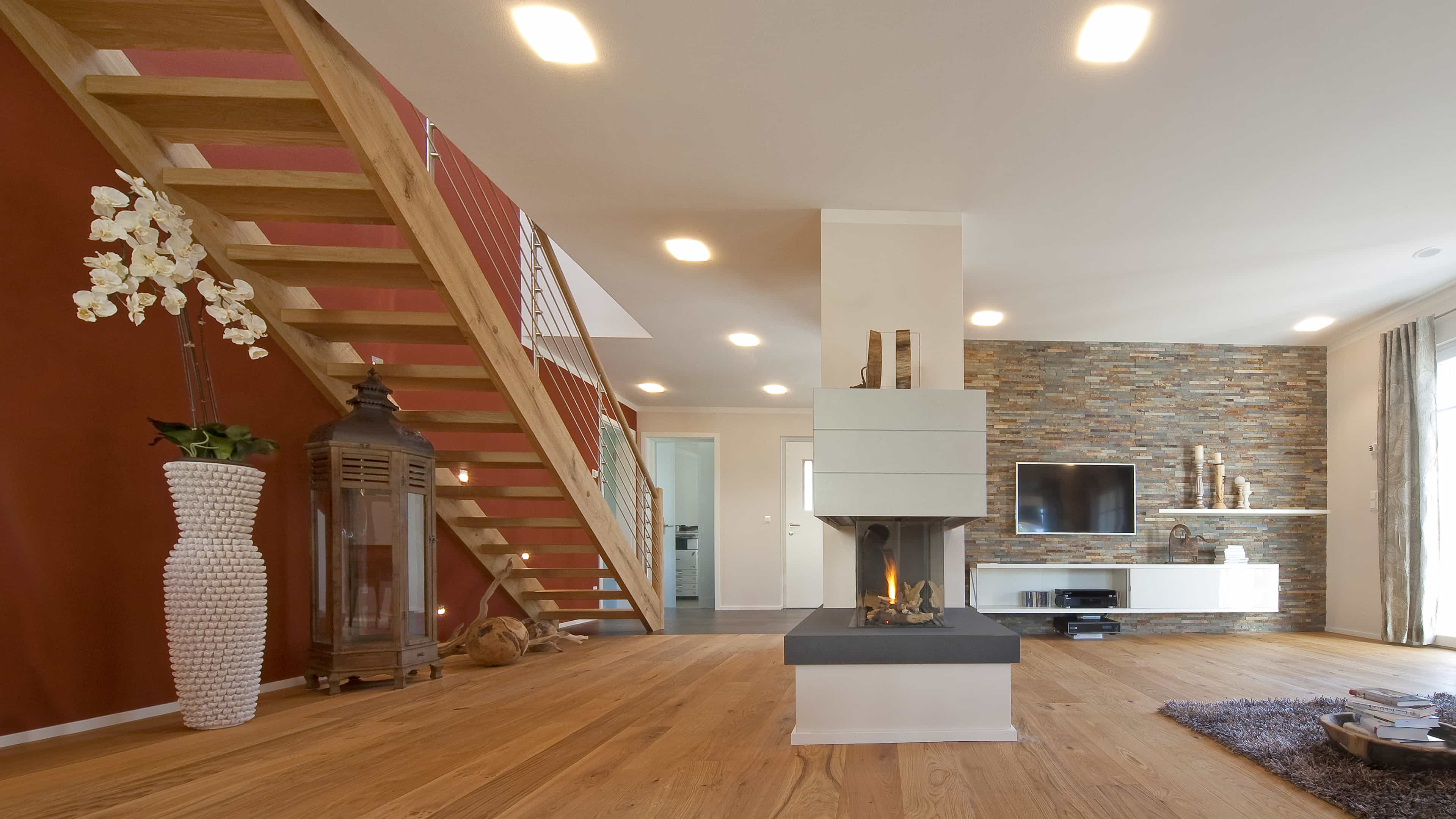 Wohnzimmer mit Kamin, Natursteinwand und offener Holztreppe im Musterhaus Haas MH Poing 187