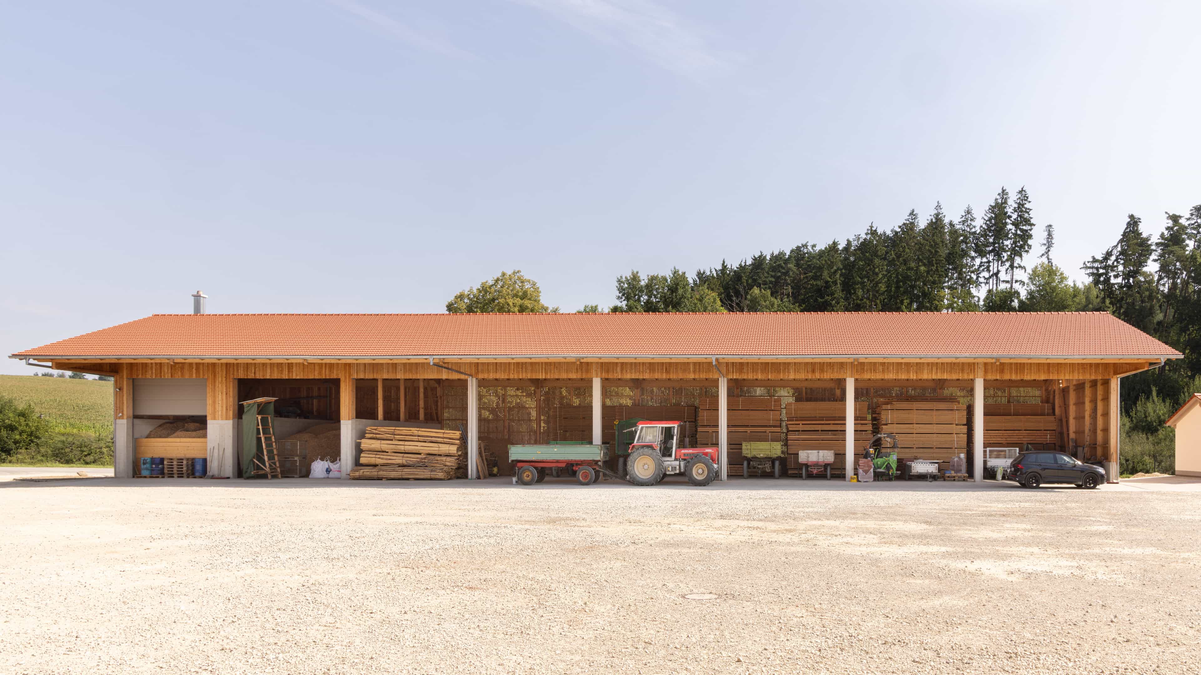 Lagergebäude mit rotem Ziegeldach, gefüllt mit Holzstapeln und landwirtschaftlichen Fahrzeugen vor einem bewaldeten Hintergrund.