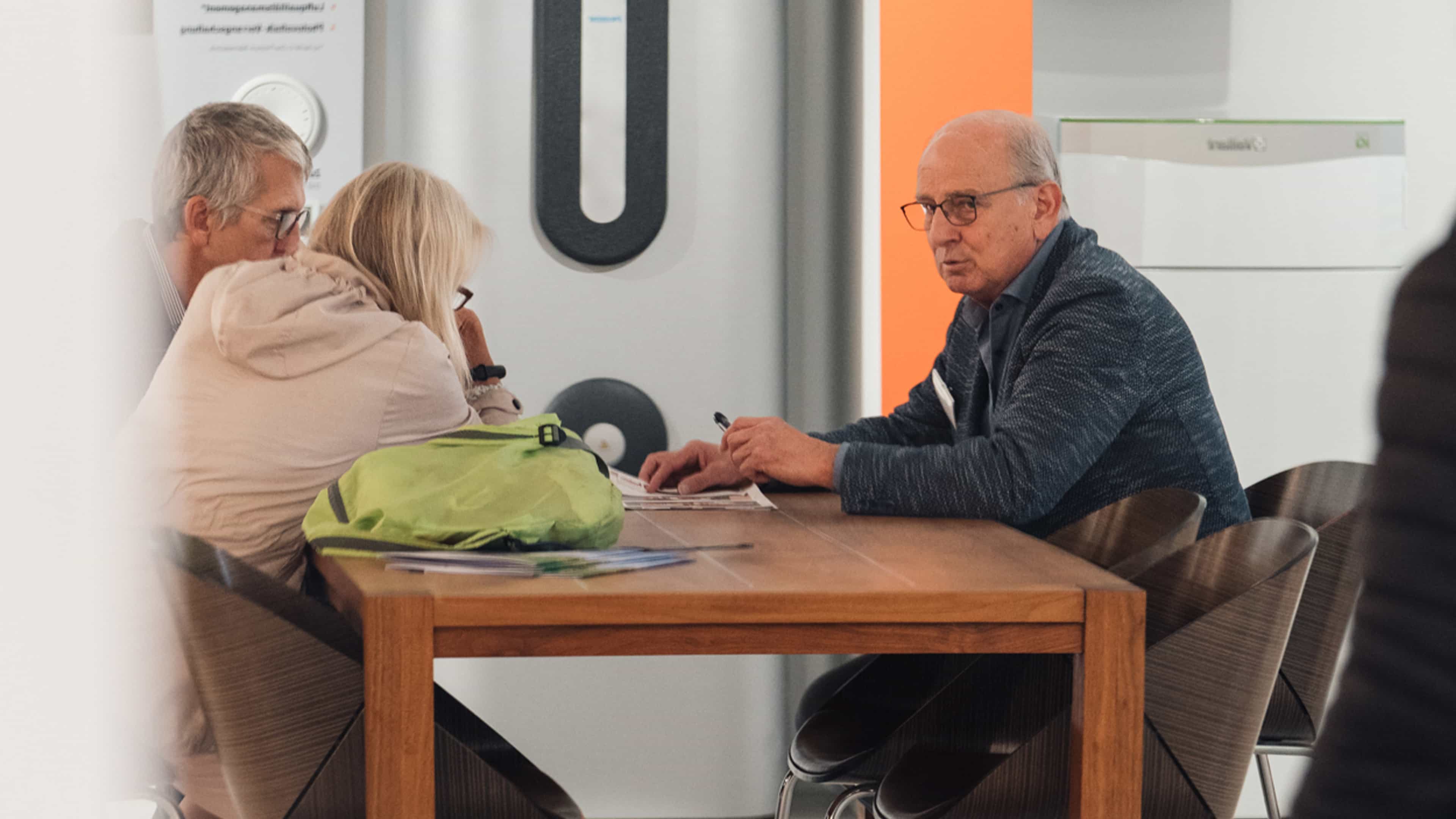 Älterer Mann berät ein Paar am Tisch, umgeben von modernen Haushaltsgeräten im Hintergrund.