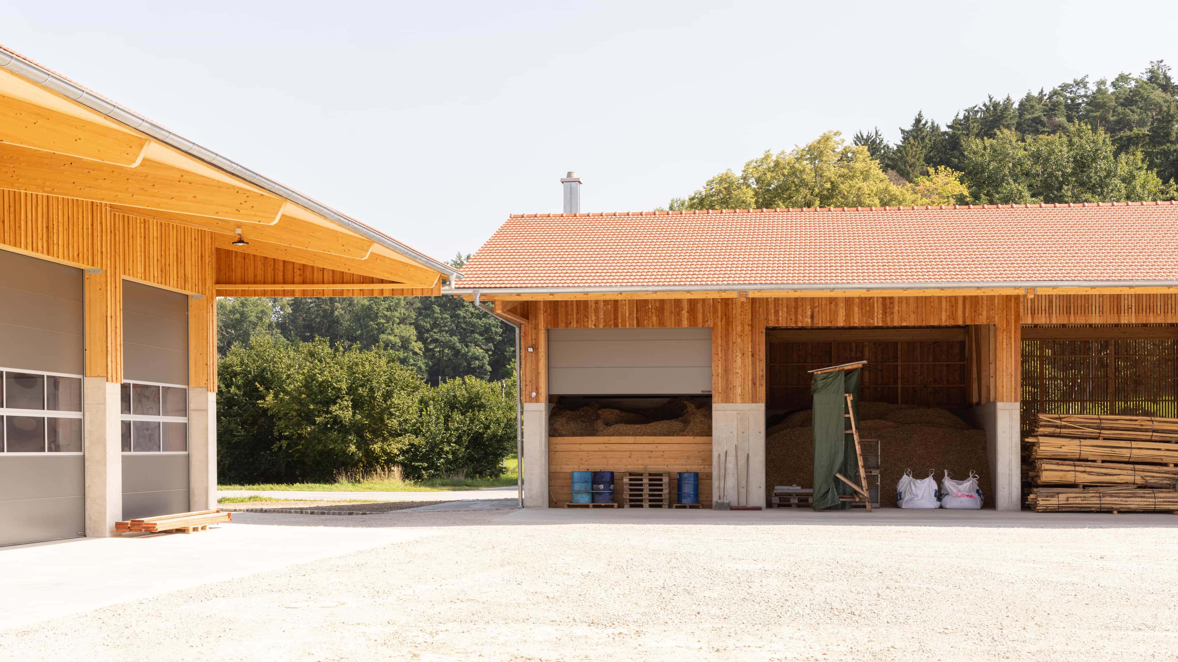Holzschuppen mit offenem Lagerraum für Holz und Pellets, umgeben von Bäumen und hellem Himmel.